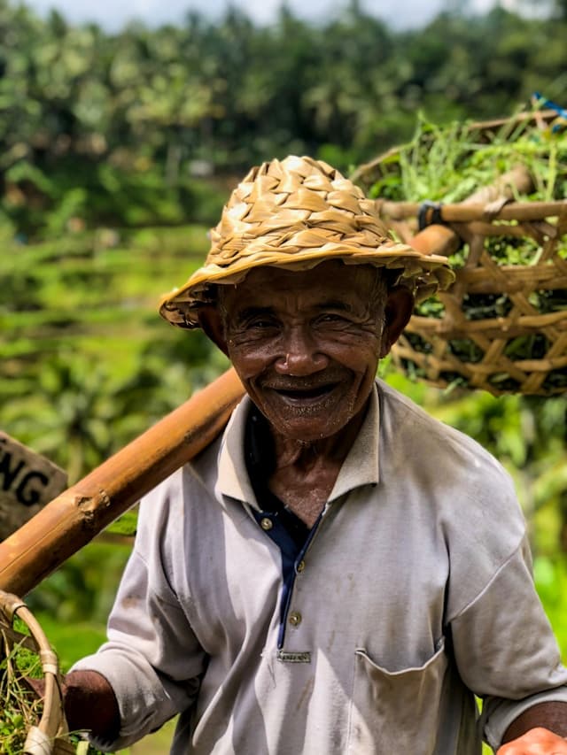 Elderly farmer in straw hat smiling while carrying a woven produce basket in terraced fields.