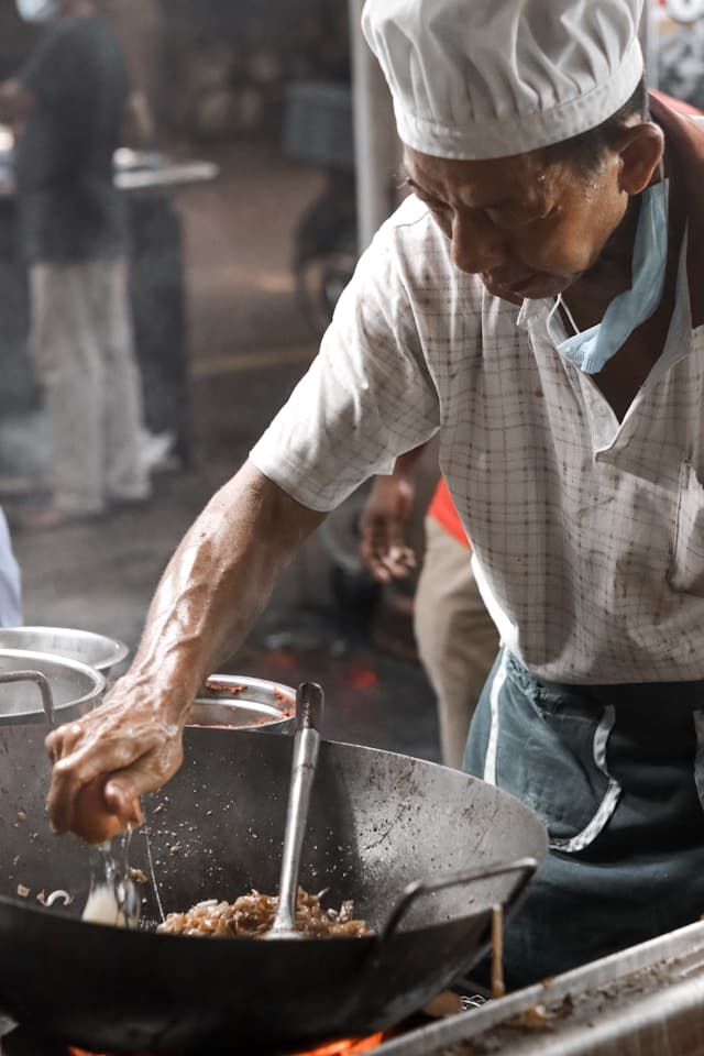 Chef in white hat stir-frying noodles over a hot wok at a bustling food stall.